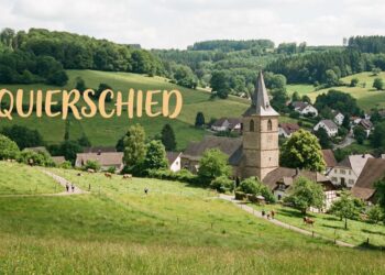 „Schwätzje Mobil" der Caritas macht Station in Quierschied und Göttelborn 2 Dorfansicht oder Landschaftspanorama der Gemeinde Quierschied mit grünen Wiesen und Waldrändern. Eventuell Kirchturm oder lokales Wahrzeichen im Mittelgrund. Der Schriftzug "QUIERSCHIED" in warmer, freundlicher Schrift, gut lesbar positioniert. Atmosphäre: Ländlich, gemeinschaftlich, naturverbunden.